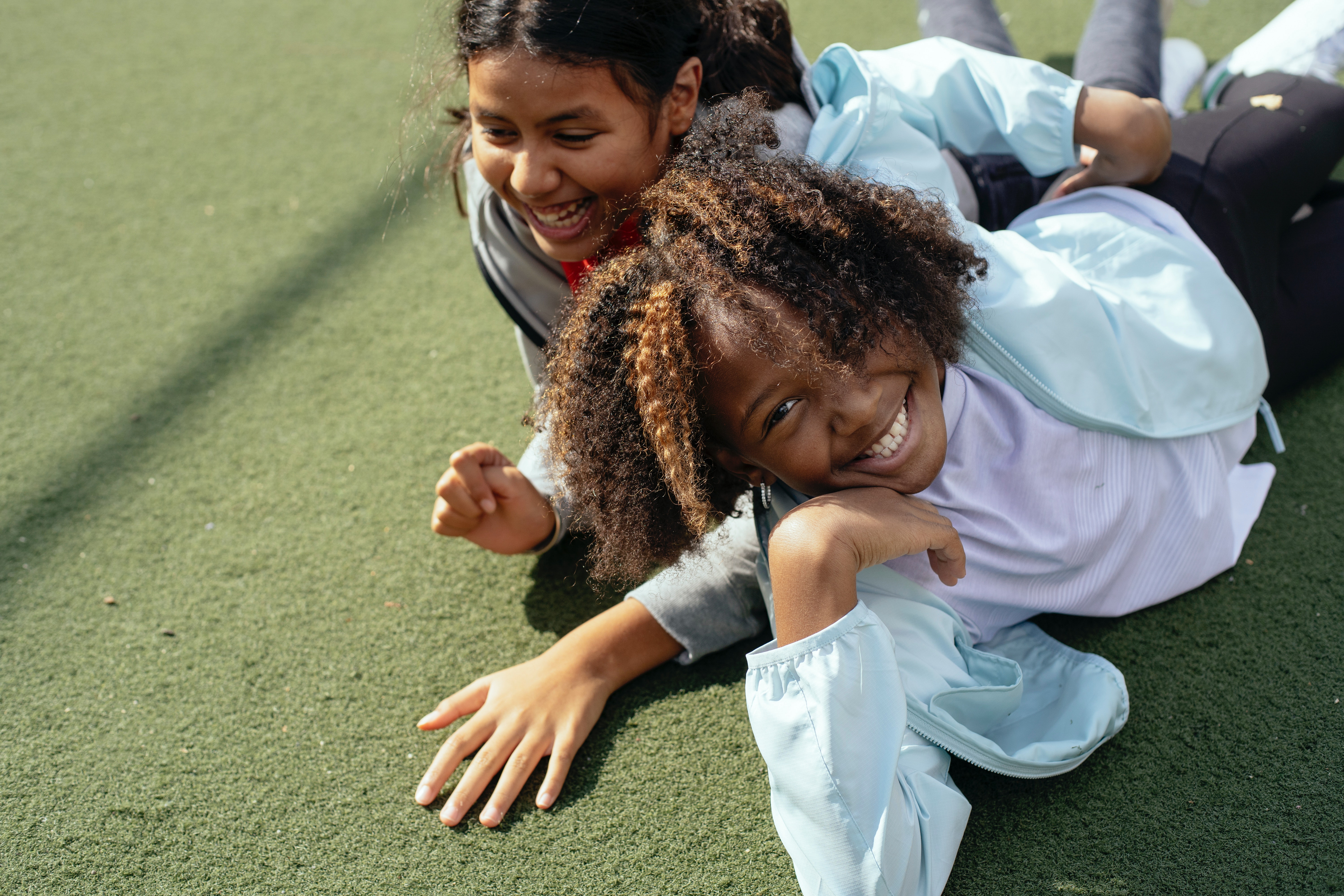 Two young girls laying side by side on a sports pitch outside. One is asian and one is black. They are both smiling.