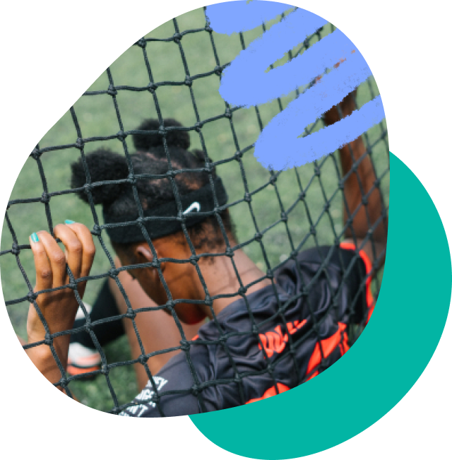 A young black girl with her back to the camera sitting down. She is in front of a sports net with her fingers interlocked with the net. She has a headband with a nike tick on it.