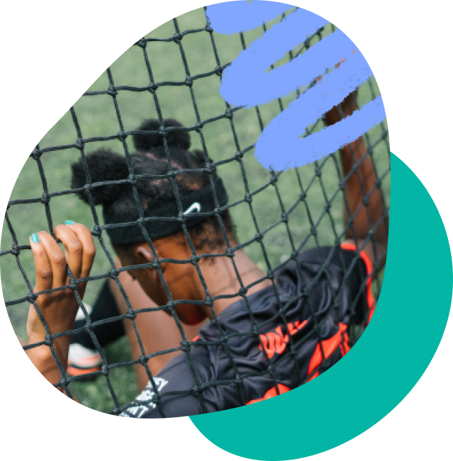 A young black girl with her back to the camera sitting down. She is in front of a sports net with her fingers interlocked with the net. She has a headband with a nike tick on it.