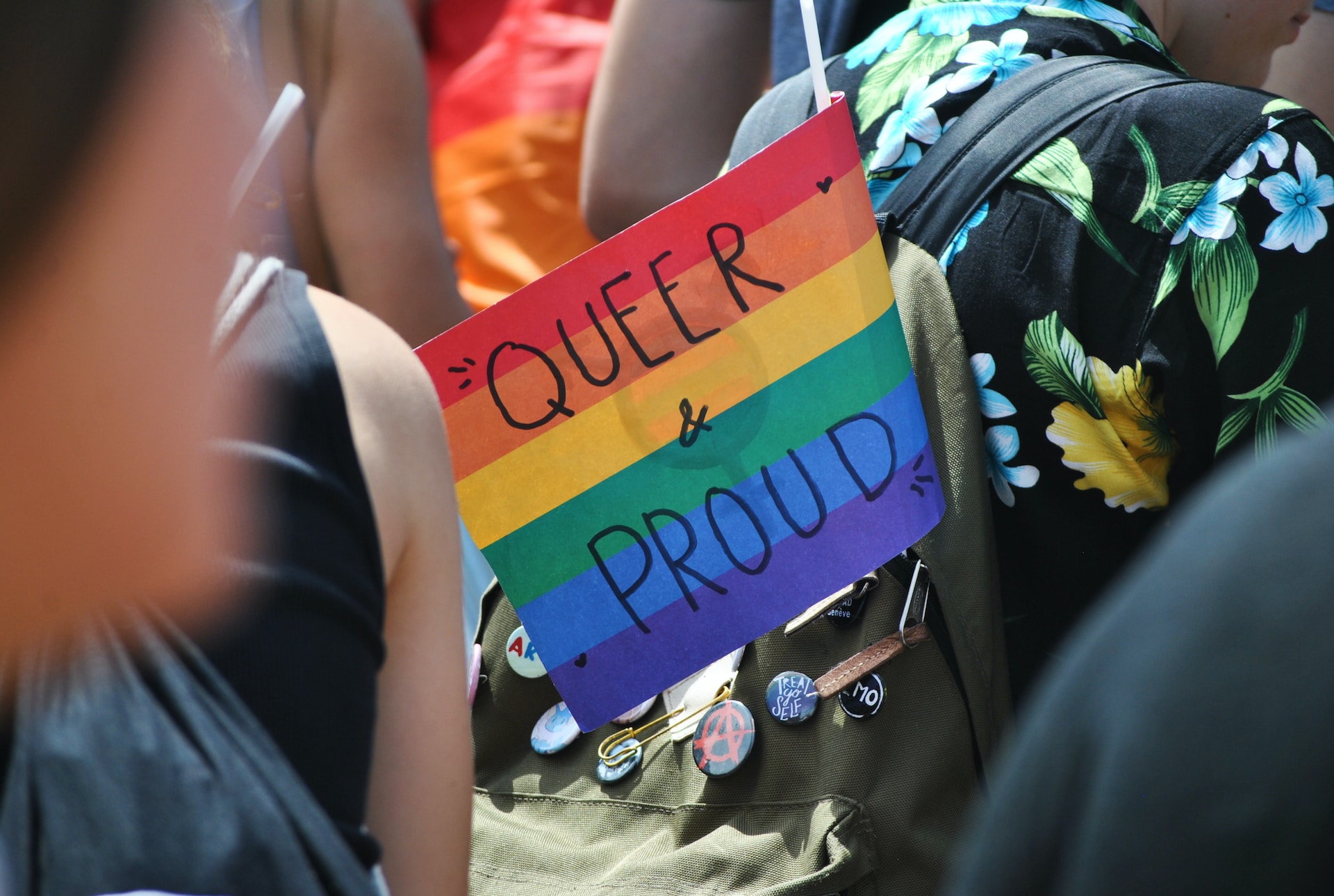 A close up of a group of people outside. You can only see one person's green khaki backpack with lots of badges on the back of it and a flag sticking out of the backpack that says queer and proud with a rainbow background.