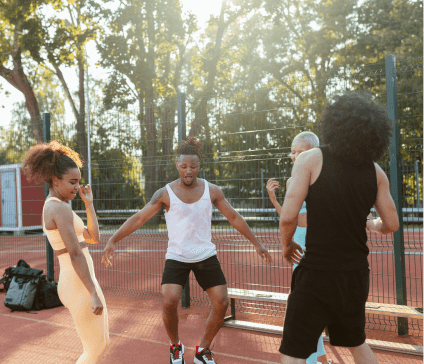 Four young mixed of different races outside on a sports pitch. They are dancing and it is sunny.