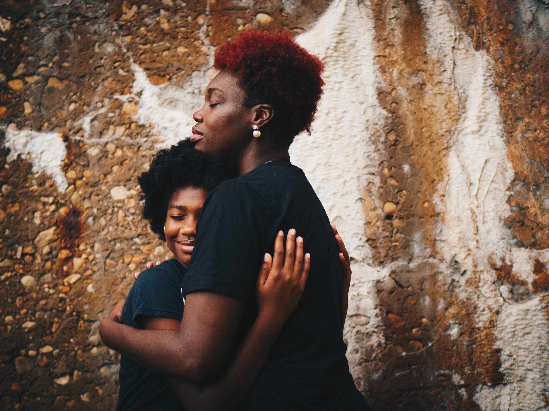 A young black girl hugging a black adult woman against a brown and white wall with stones in it. The young girl is smiling. They both have their eyes closed. The black adult woman has red hair and pearl earrings.