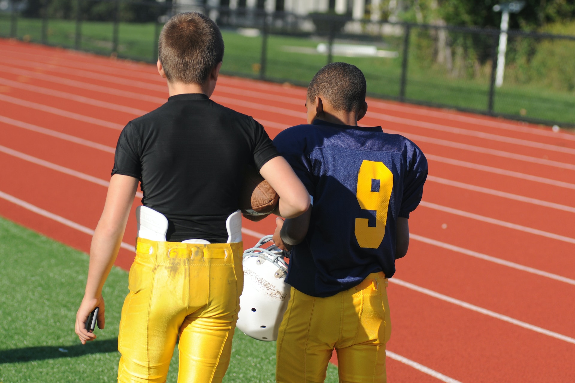 Two young boys are wearing football kit and walking away from the camera and on a football pitch. One is white and one is black. One has a phone in their hand and a football. The other is holding their football helmet and is wearing a blue shirt with the number nine on it.