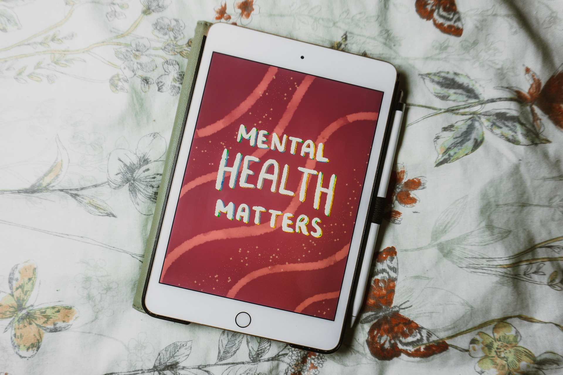 A close up of a white iPad and pen with the words mental health matters on the screen on a background with some squiggles. The iPad is on a bed with a cover that is white with red, yellow and green butterflies and flowers.
