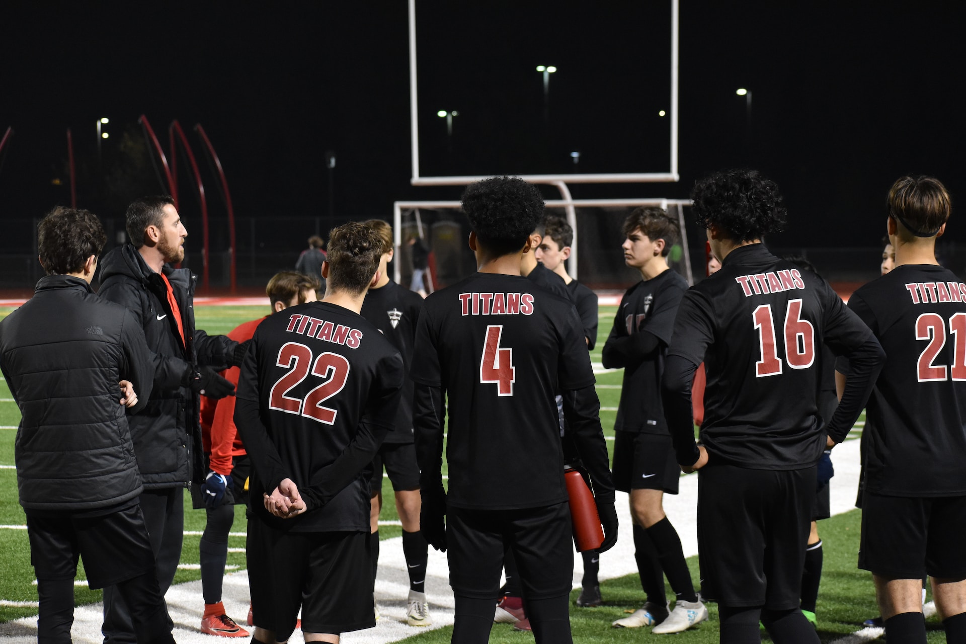 A male coach talking to his team on a sports field. It is night time and the field is lit with flood lights. The team are all young males of different races wearing black team kit and TITANS on the back of their shirts.
