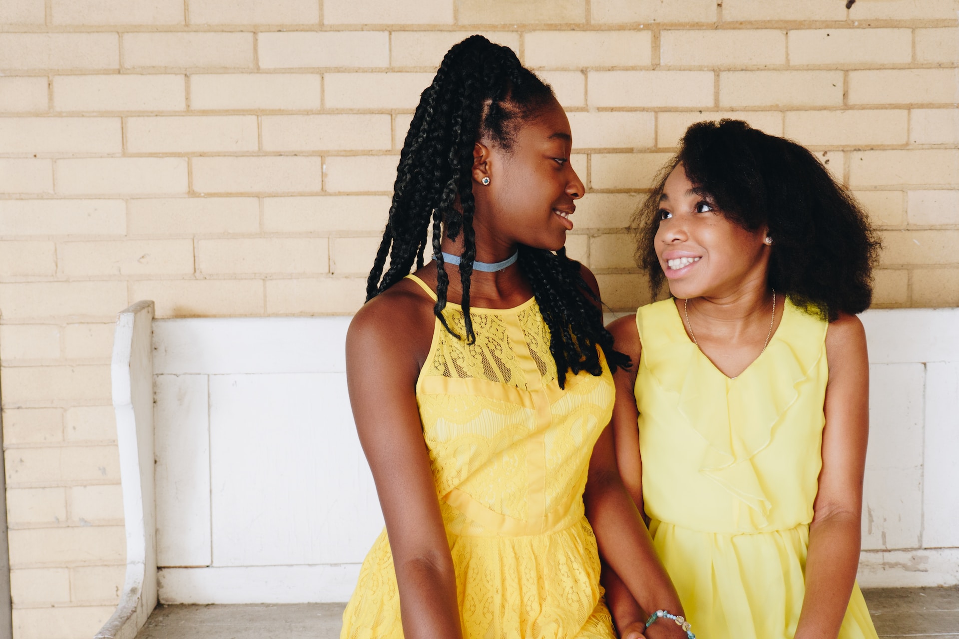 Two young black girls sitting side by side wearing yellow dresses. They are on a white bench and are looking into each others eyes smiling and holding hands.
