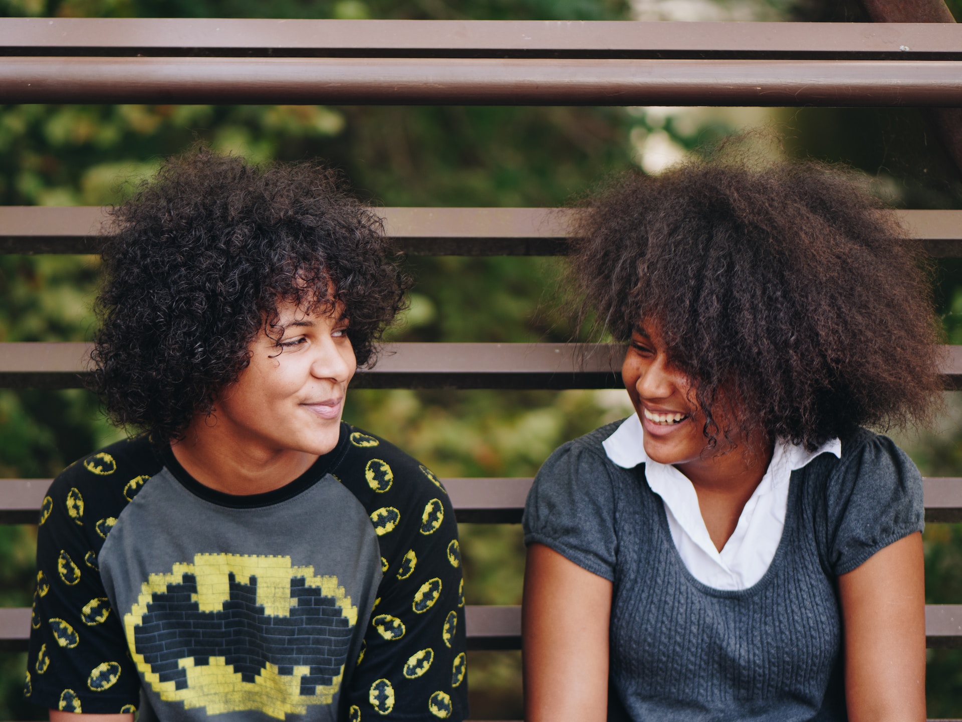 Two young black people sitting on a bench outdoors looking toward each other and smiling. One is wearing a grey top over a white shirt, the other is wearing a t-shirt with repeated batman symbols.