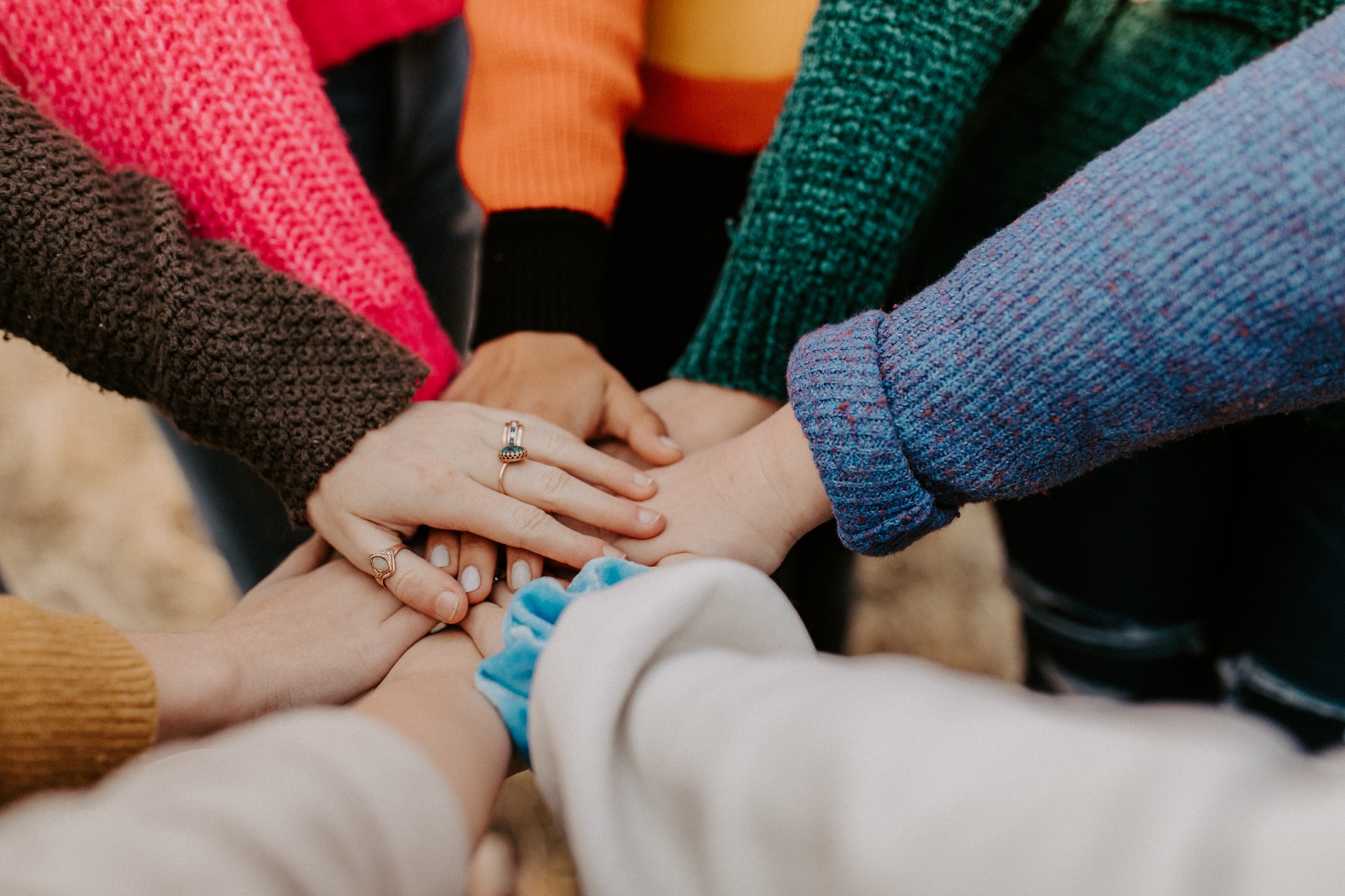 A group of young people with their hands together and a close up of their hands. They are wearing different colored sweaters and the hand on the top has three rings.