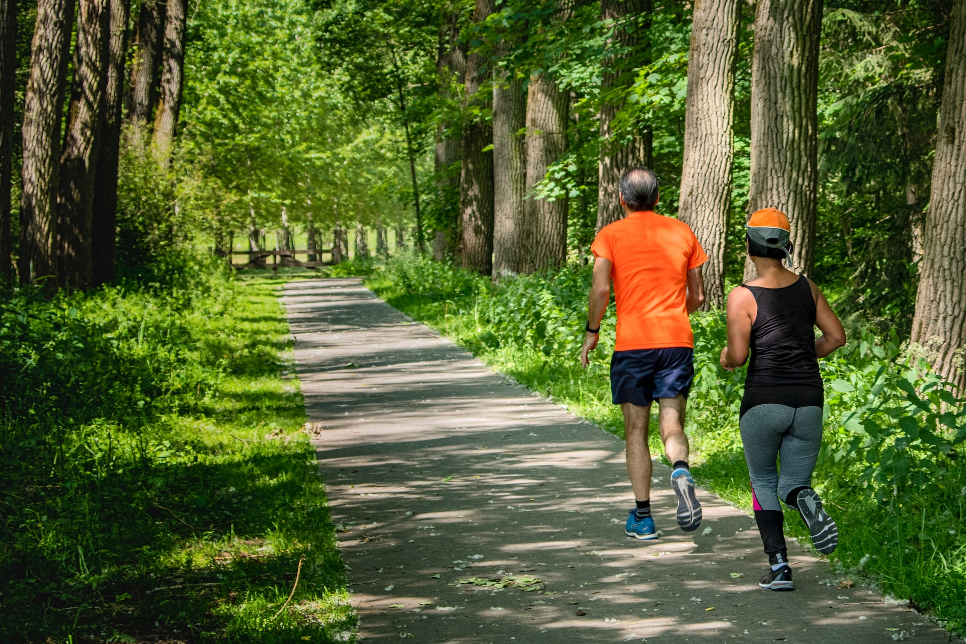 A man and a woman running along a concrete path in a lush green park with trees either side.