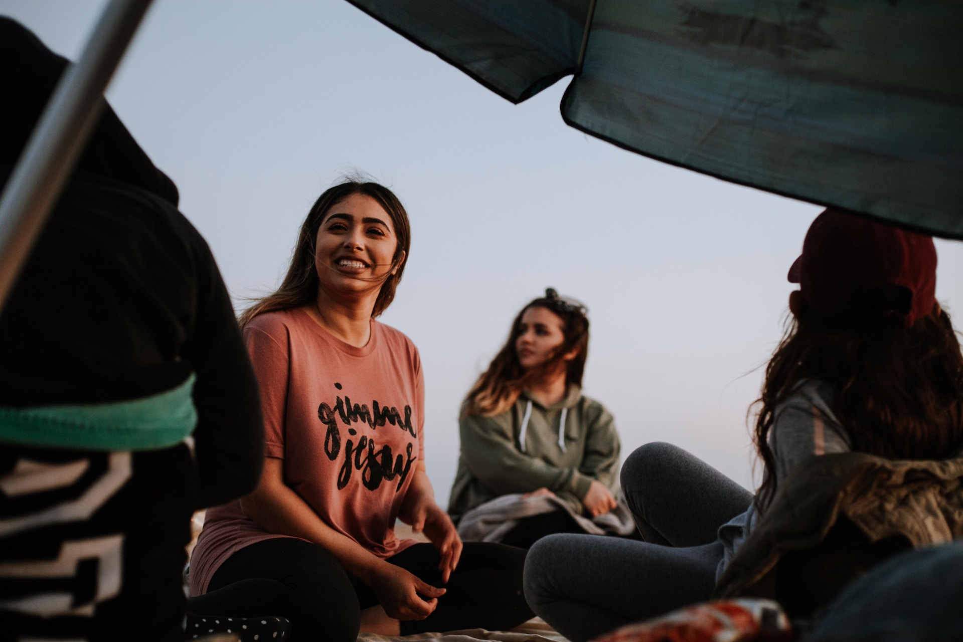 A group of young people camping outdoors. One girl is smiling at her friend. Another girl is looking into the distance. Two others have their backs to the camera. They are surrounded by blankets and cushions.