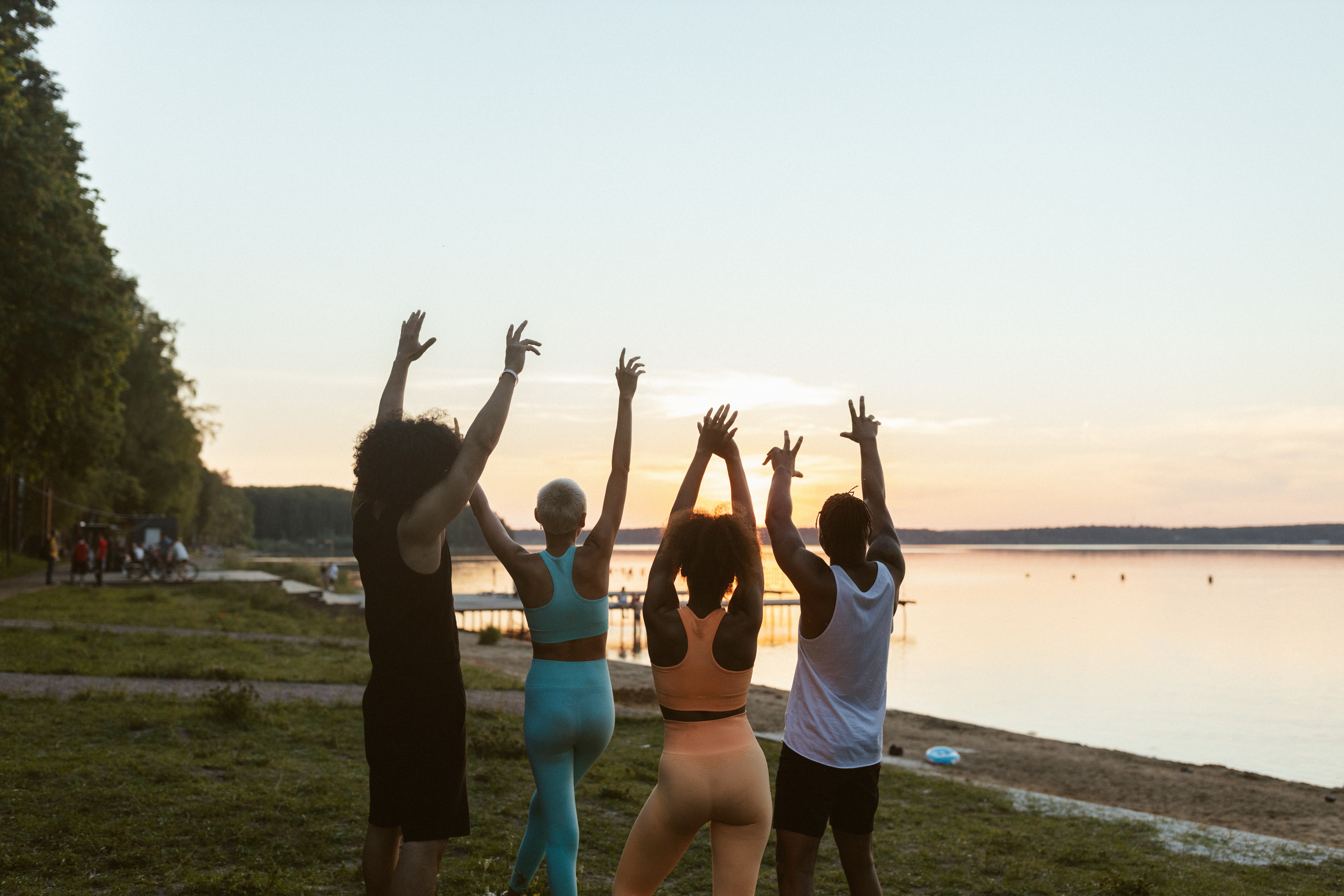 Four young people on grass at sunset looking out over a lake. They have their arms up in the air with their backs to the camera.