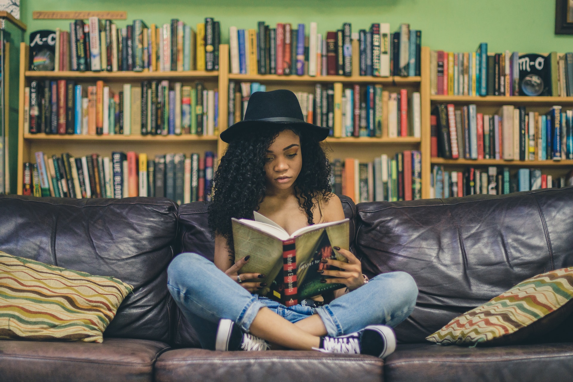 A young mixed race woman sitting on a sofa of a library with her legs crossed reading a book. She has a hat on and there are shelves full of books behind her.
