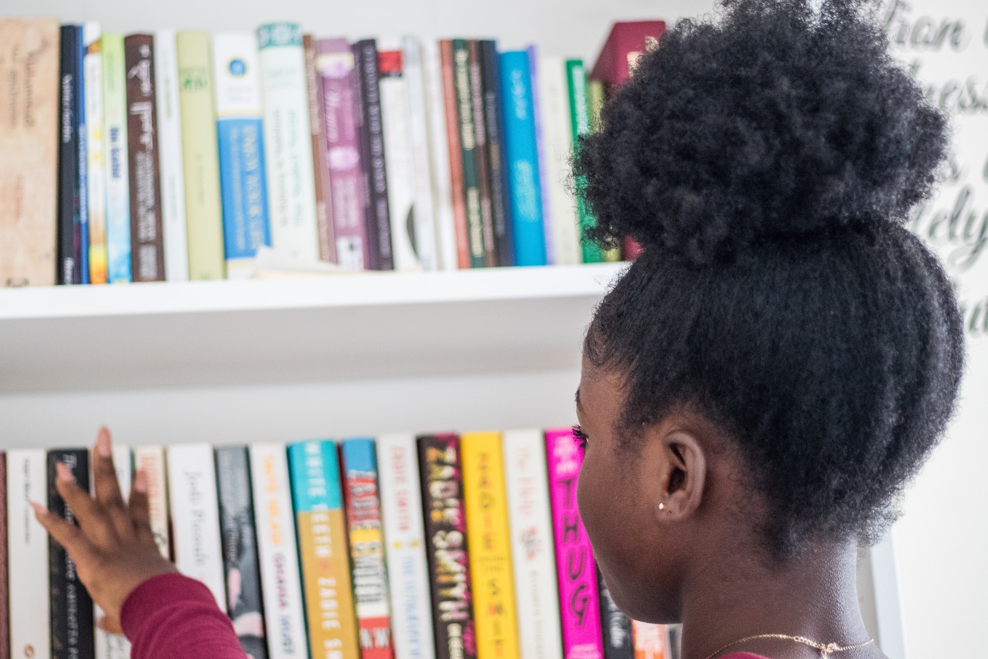 A young black girl is looking across a row of books on a book shelf.